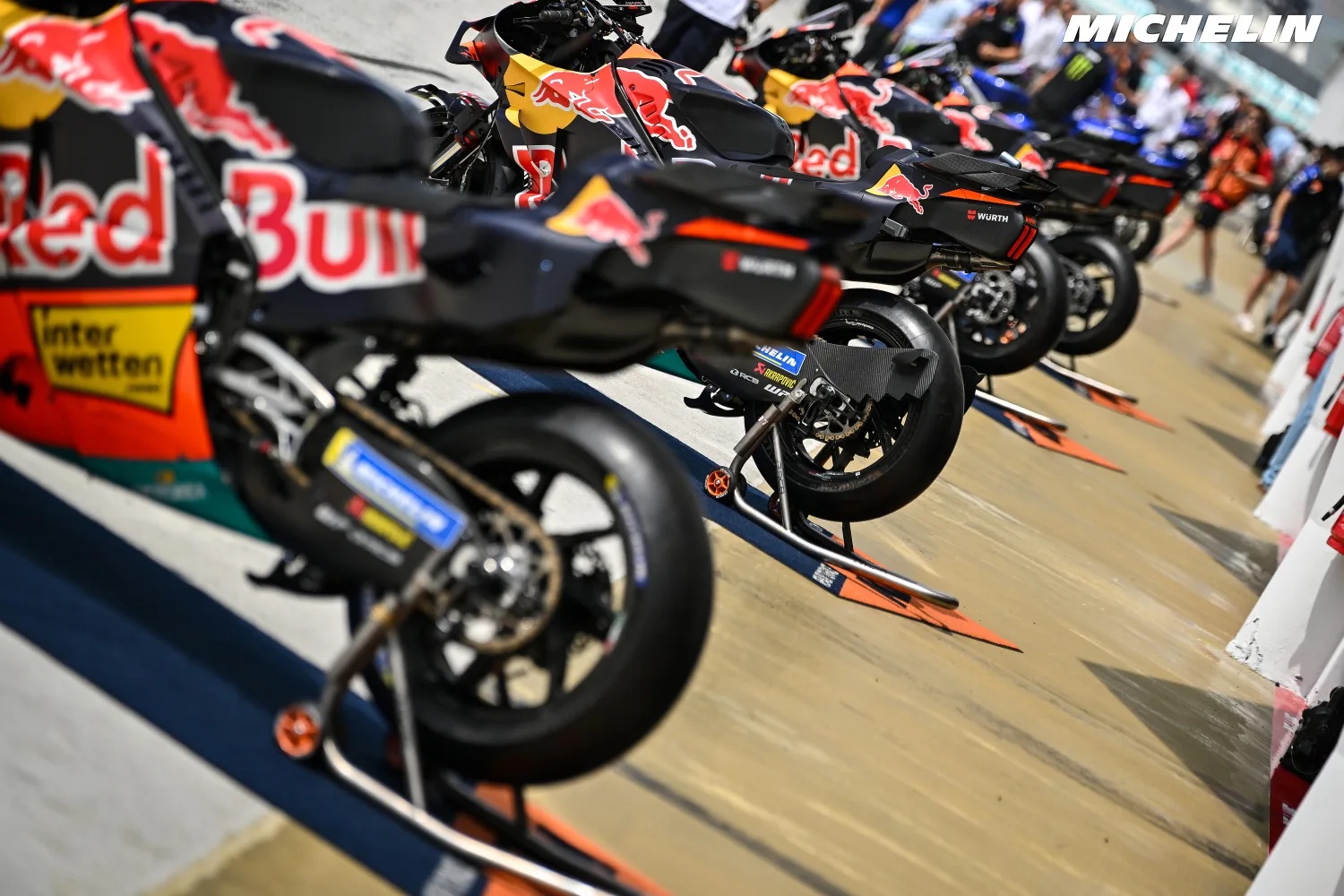 Row of Red Bull-liveried MotoGP bikes on stands in the paddock, with Michelin branding visible, as riders gather nearby.