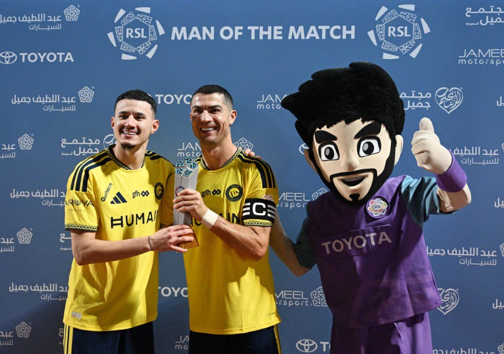 Cristiano Ronaldo and a teammate hold a Man of the Match trophy next to a mascot in front of a branded backdrop.
