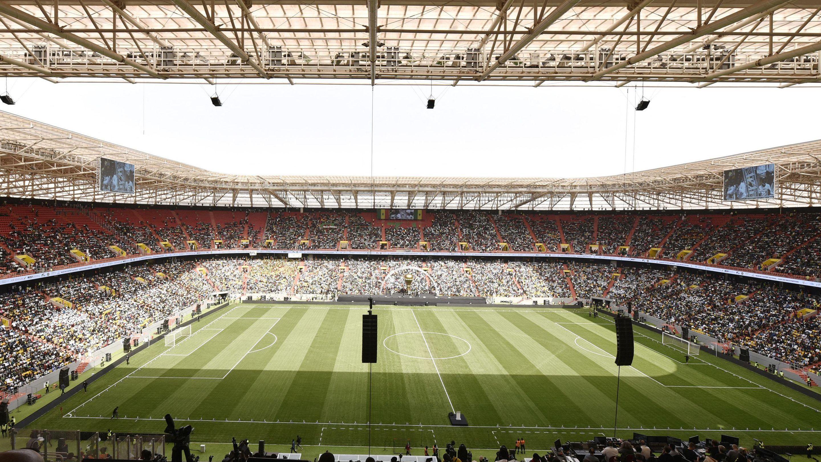 An interior view of the Abdoulaye Wade Stadium taken from the top tier showing a grass football pitch and packed stands. Video screens can be seen suspended from the white roof above the football goals at either end