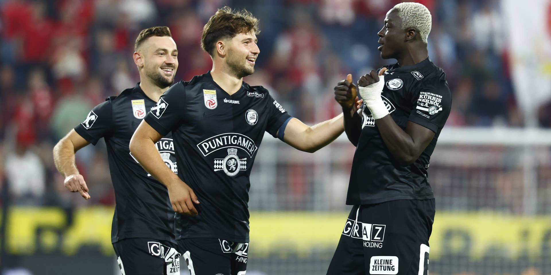 Drei Fußballer des SK Sturm Graz jubeln in schwarzen Trikots gemeinsam auf dem Spielfeld.