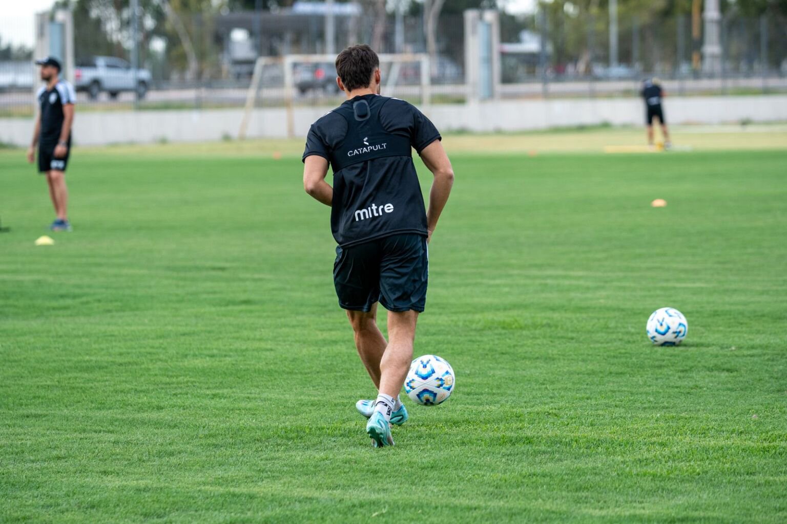 A footballer in black training kit runs with the ball across an empty training pitch