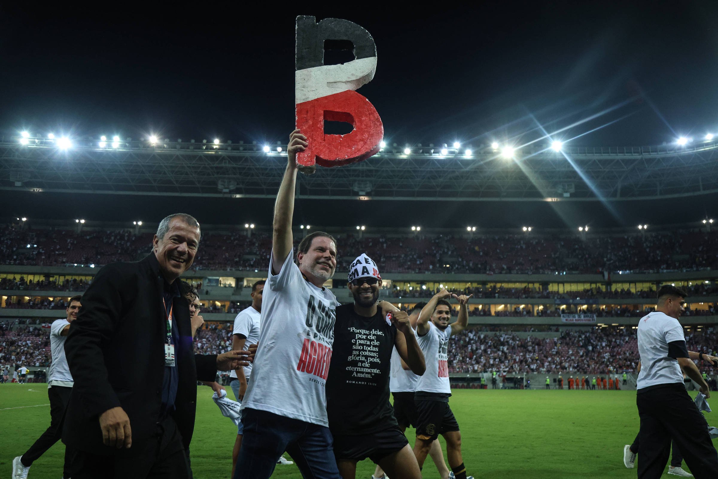 Homens no campo de um estádio de futebol à noite, um deles segurando uma letra 'B' pintada nas cores preto, branco e vermelho. Público nas arquibancadas ao fundo e iluminação forte no estádio.