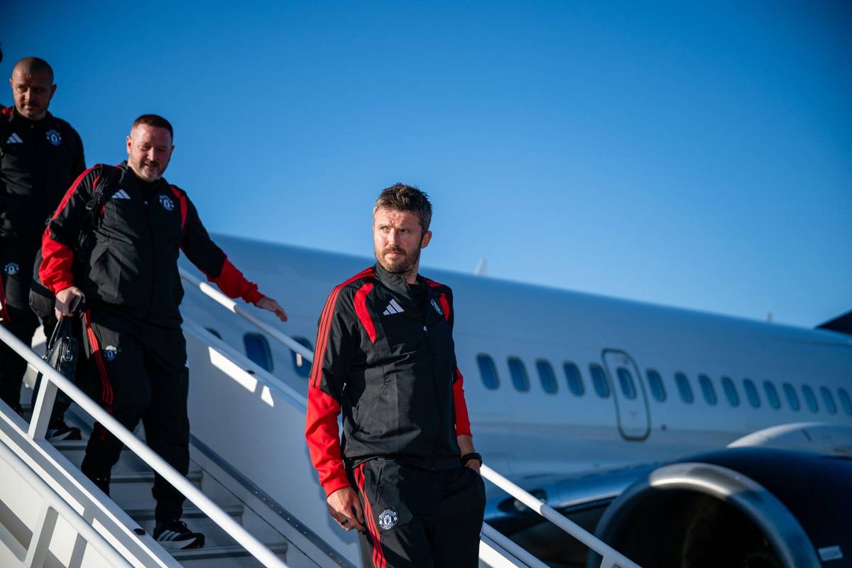 An individual dressed in a black and red Adidas sports jacket, standing in an outdoor setting with a clear blue sky in the background.