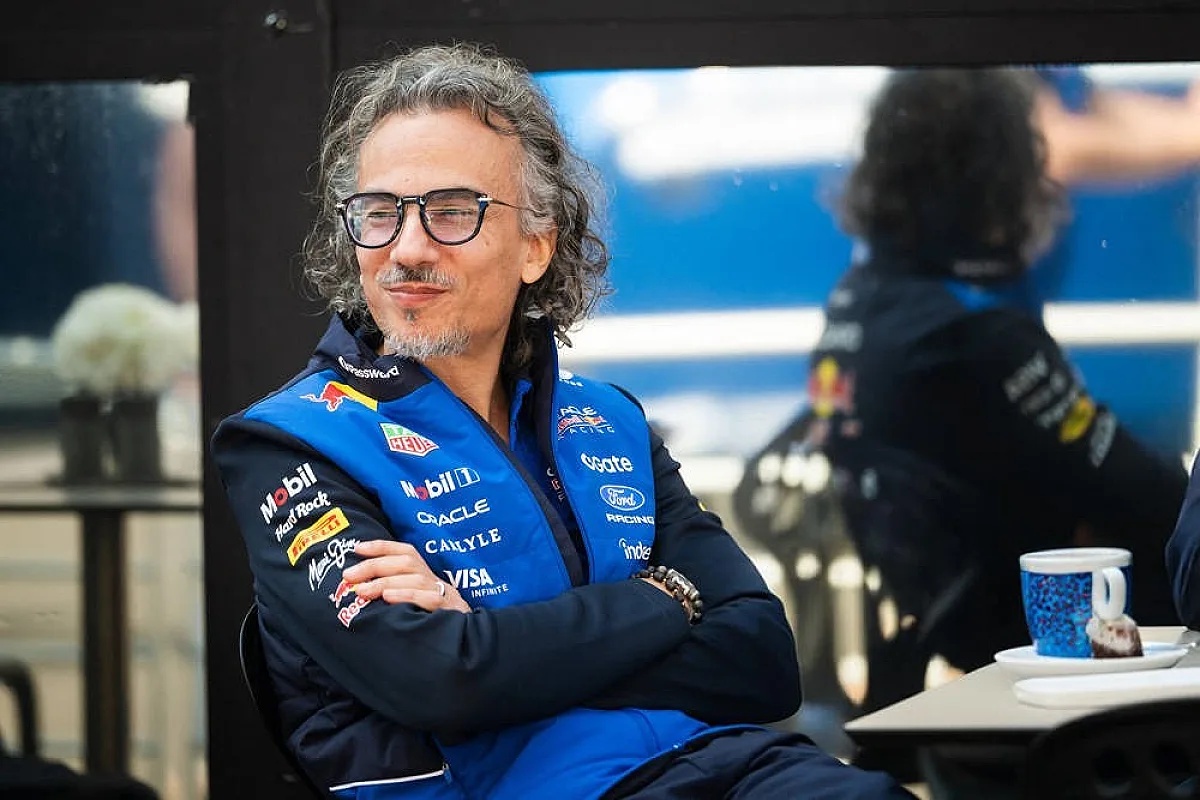 Man with curly grey hair and glasses, wearing a navy and blue Red Bull team jacket covered in sponsor logos, arms crossed at a paddock table.