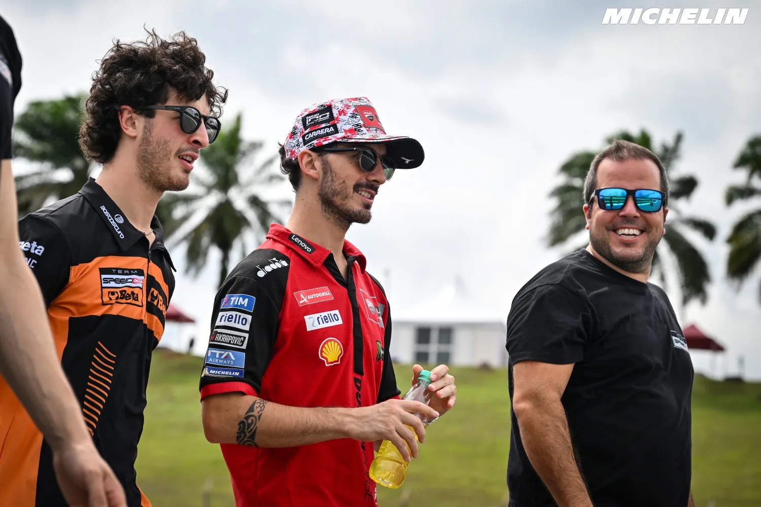 Three men walk through a paddock in MotoGP-style team gear; one wears a red Ducati-branded shirt with Shell and Ducati logos and a patterned cap.