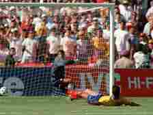 Colombian defender Andres Escobar lies on the ground in front of his own goal as goalkeeper Oscar Cordoba reacts, with the ball entering the net and a crowd watching behind them.