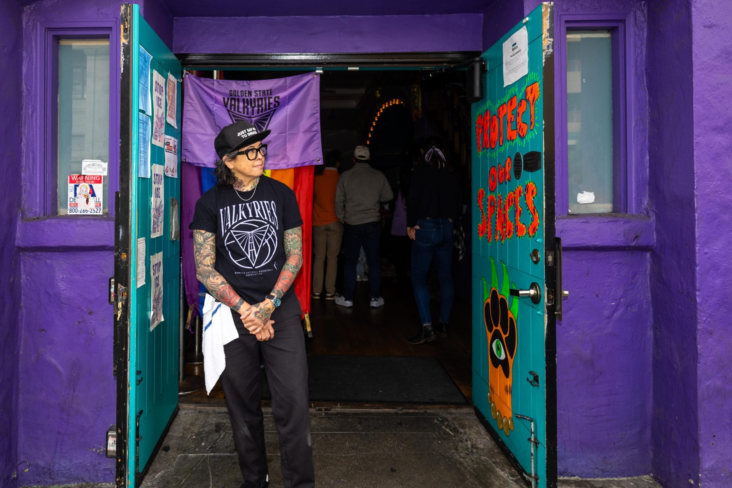 A person wearing a Golden State Valkyries t-shirt and hat stands in the doorway of a purple building with a “Protect Our Spaces” sign on the door.