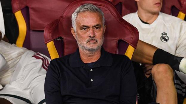 Jose Mourinho sits, straight-faced, on a seat in dark clothing, with Real Madrid players in white kit sitting behind him