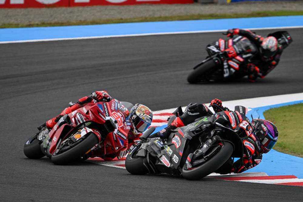 MotoGP riders in red Ducati and dark Aprilia-style liveries racing through a corner on a track with red-and-blue kerbs