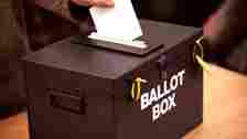 BBC A black box which reads Ballot Box is on a table, someone is posting a ballot paper through the hole in the top of it