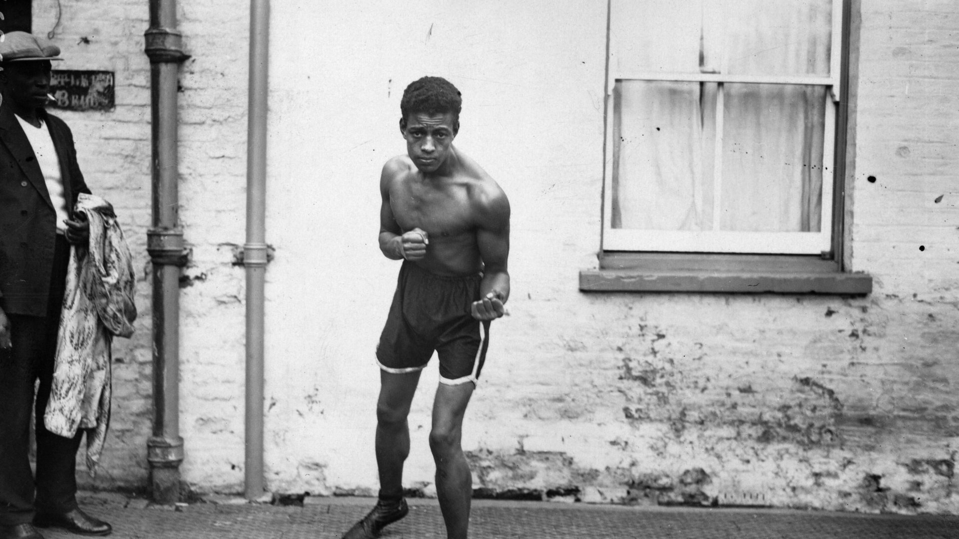 Johnson stands in boxing shorts and pumps with his fists clenched looking at the camera.
