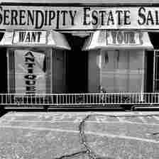 A black-and-white photo of a storefront titled "Serendipity Estate Sales." The left window displays the word "WANT" above a sign that reads "ANTIQUE." The right window displays the word "YOUR." The sidewalk in front appears weathered with visible cracks.