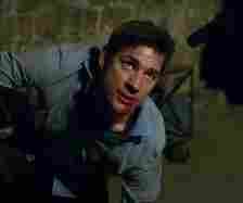 John Krasinski, disheveled and injured, is on his knees with blood on his face, looking up in a tense moment.