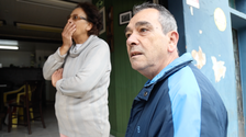 João Batista Coelho, in a blue sports jacket, stands outside his store, while his wife looks on, a hand over her mouth, as if overcome with emotion.