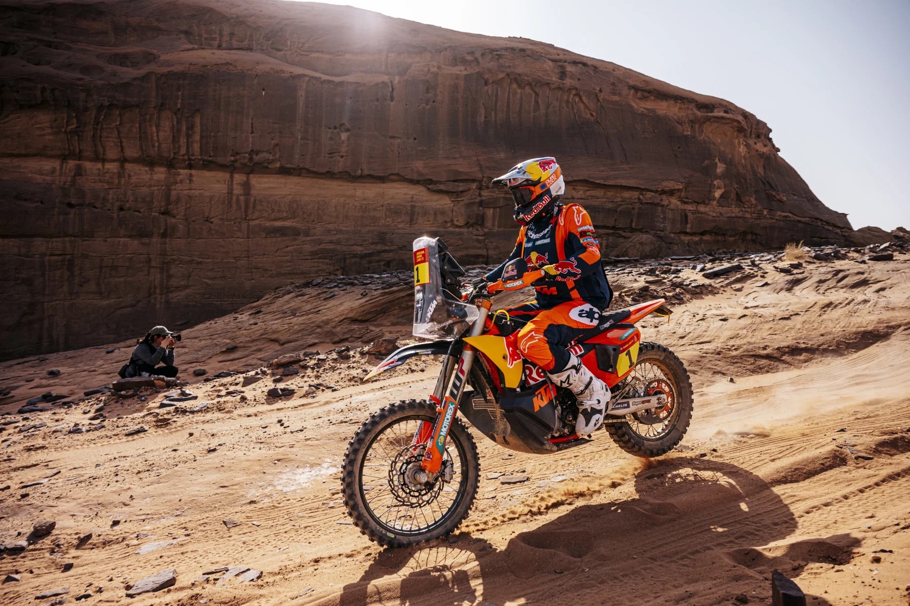 Rider in orange KTM Red Bull leathers and helmet on a KTM Rally bike, racing through sandy desert terrain near red rock cliffs at the Dakar Rally