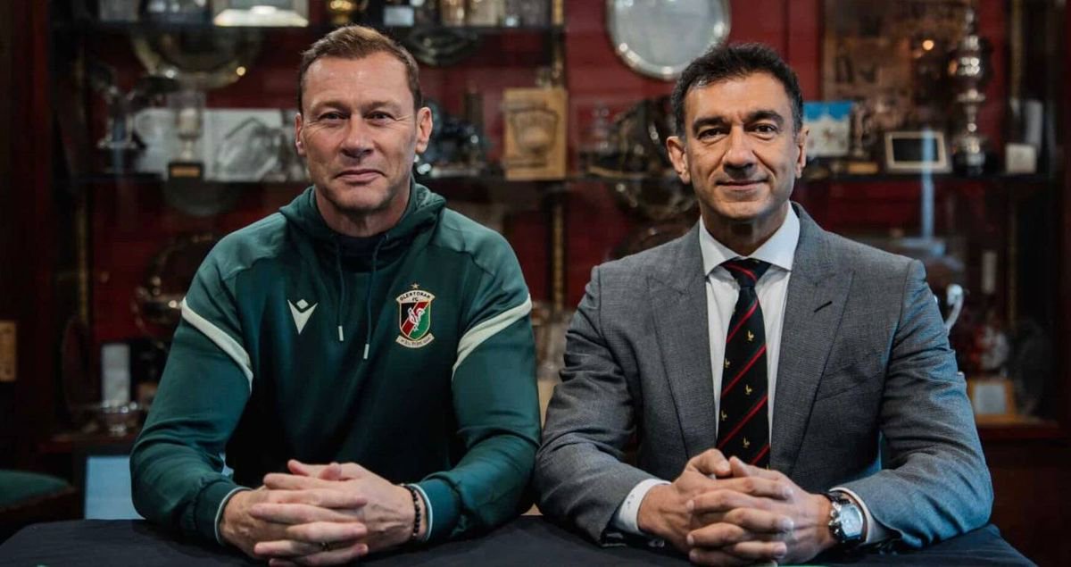 Two men are seated at a table, likely in a formal setting, with one individual wearing a green jacket and the other in a gray suit and tie. They appear to be engaged in a discussion or interview, with various decorative items and a wall in the background.