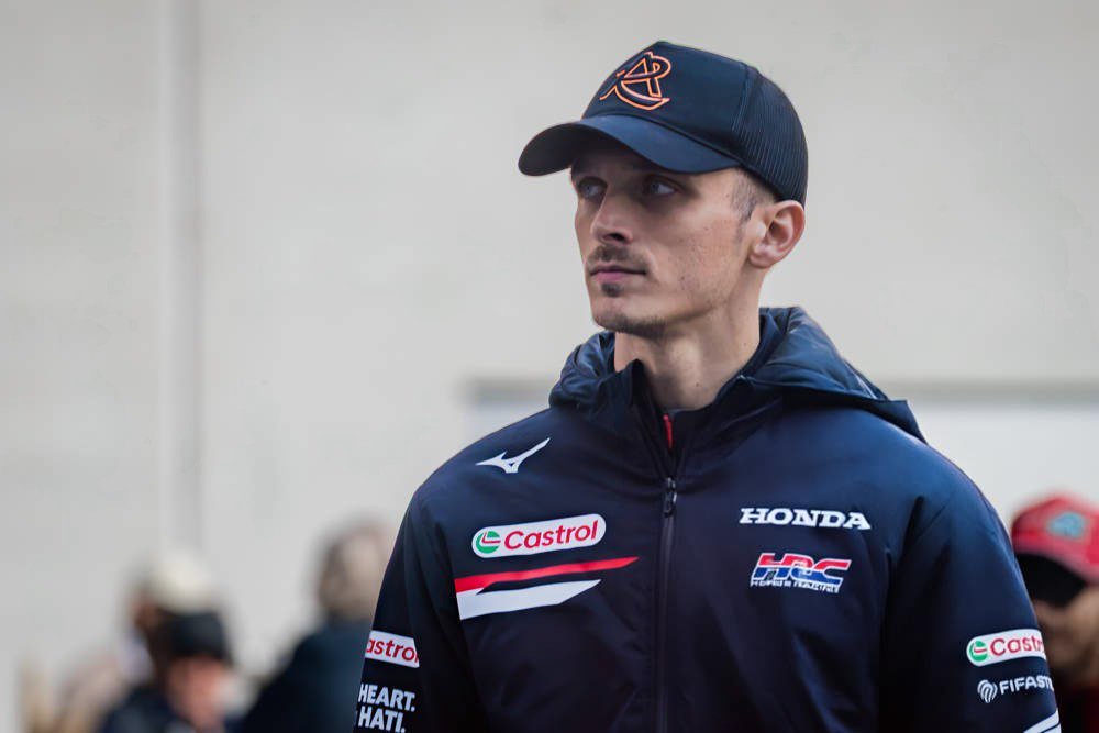 A rider in a navy Honda HRC team jacket with Castrol and Mizuno logos and a dark cap, standing in the MotoGP paddock