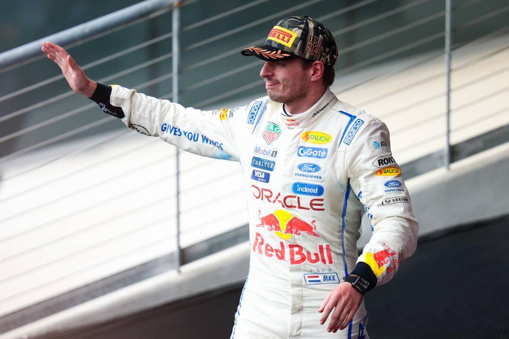 Driver in white Red Bull Racing suit with Oracle and Red Bull logos, cap with Pirelli logo, waving in the paddock; article about Max Verstappen