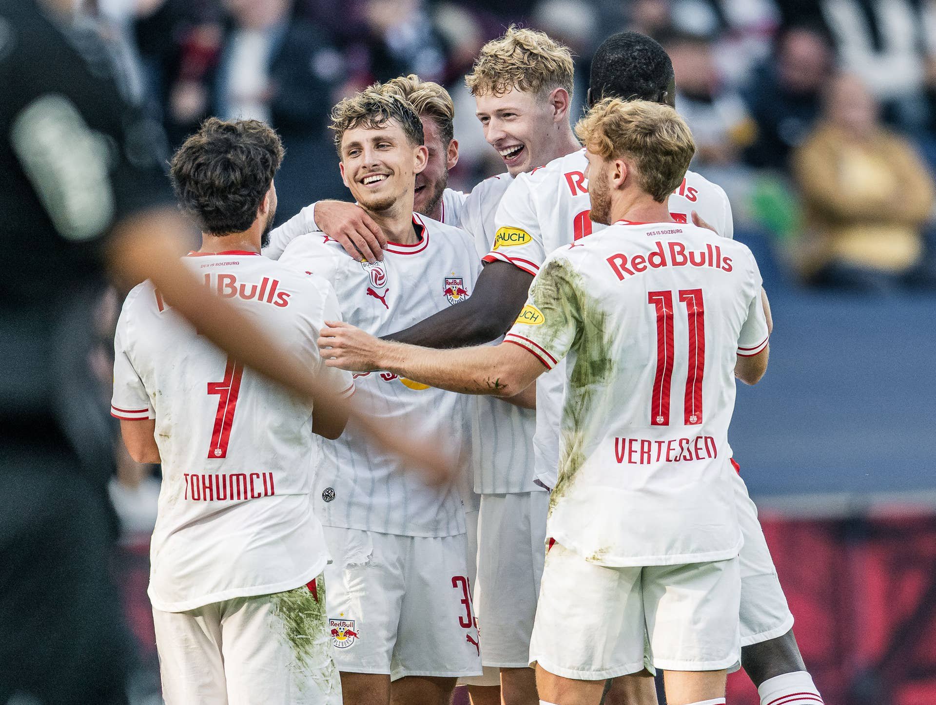 Mehrere Spieler von Red Bull Salzburg in weißen Trikots umarmen sich strahlend auf dem Spielfeld und feiern gemeinsam ein Tor.