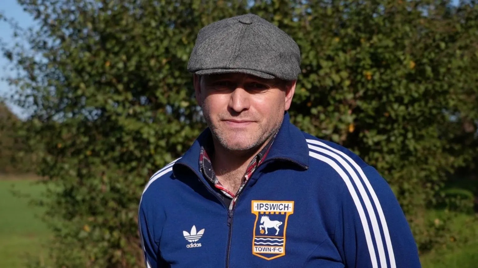 BBC Aaron Rose is wearing a flat cap and a blue Adidas zip-up jacket with white stripes on the arms and an Ipswich Town badge on his chest. He is standing in a grassed area, with hedging behind him,