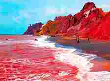 Picture of surf rolling up on sand with water a red color and red-tinted rocks rising behind the beach