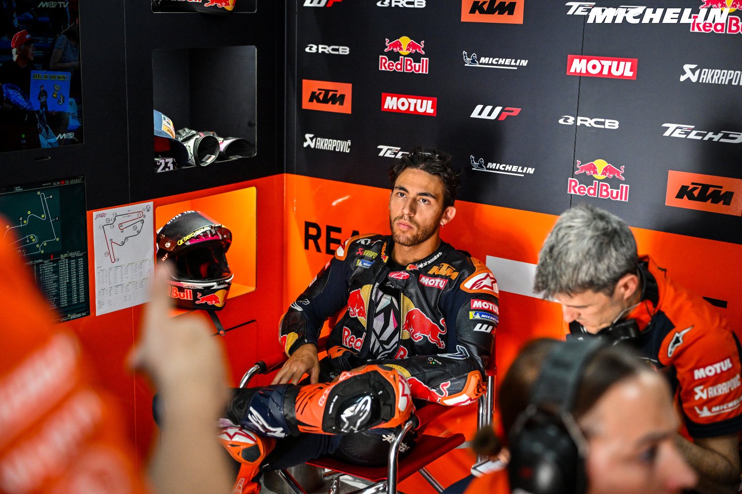 A rider in KTM Red Bull racing leathers sits in the team garage, with KTM and Red Bull branding behind him and a helmet nearby