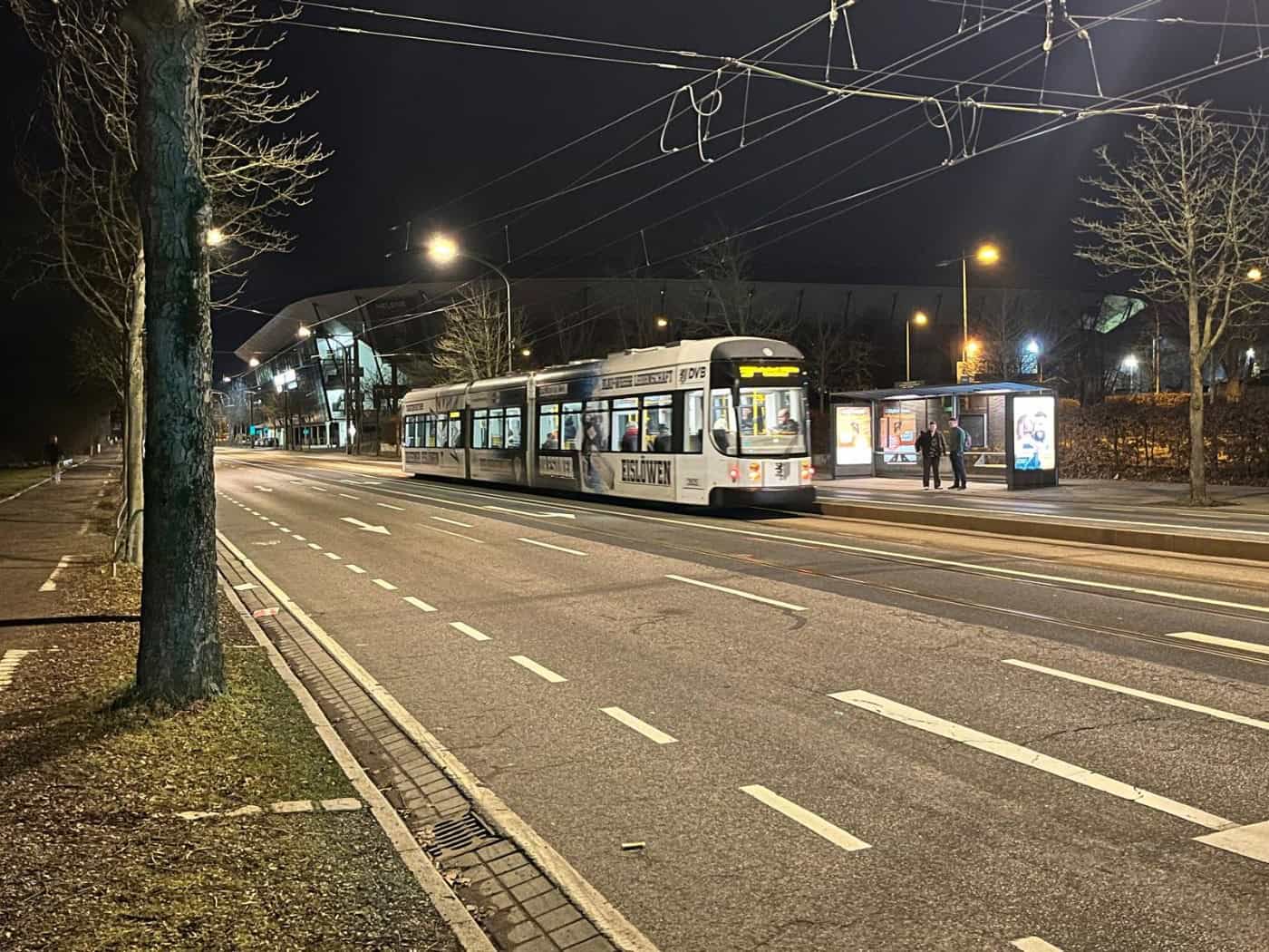 Nachtaufnahme einer beleuchteten Straßenbahn der Dresdner Verkehrsbetriebe an einer Haltestelle vor der dunklen Kulisse des Rudolf-Harbig-Stadions. Straßenlaternen erhellen die mehrspurige, asphaltierte Straße im Vordergrund.