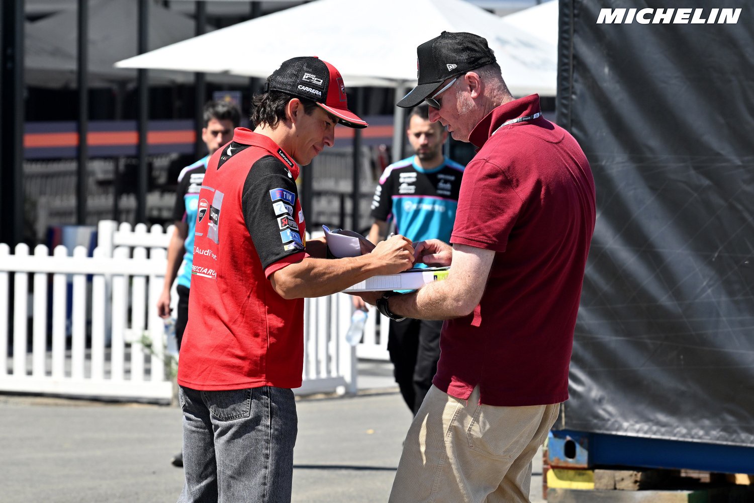 A rider in a red cap and red vest signs an autograph for a fan in a MotoGP paddock area, with team staff and a Michelin banner behind them