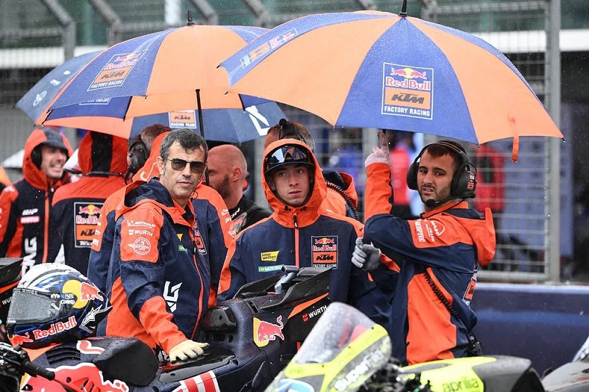 Red Bull KTM Factory Racing crew in orange and navy rain gear under umbrellas at a wet MotoGP grid, one rider in a helmet beside a KTM bike