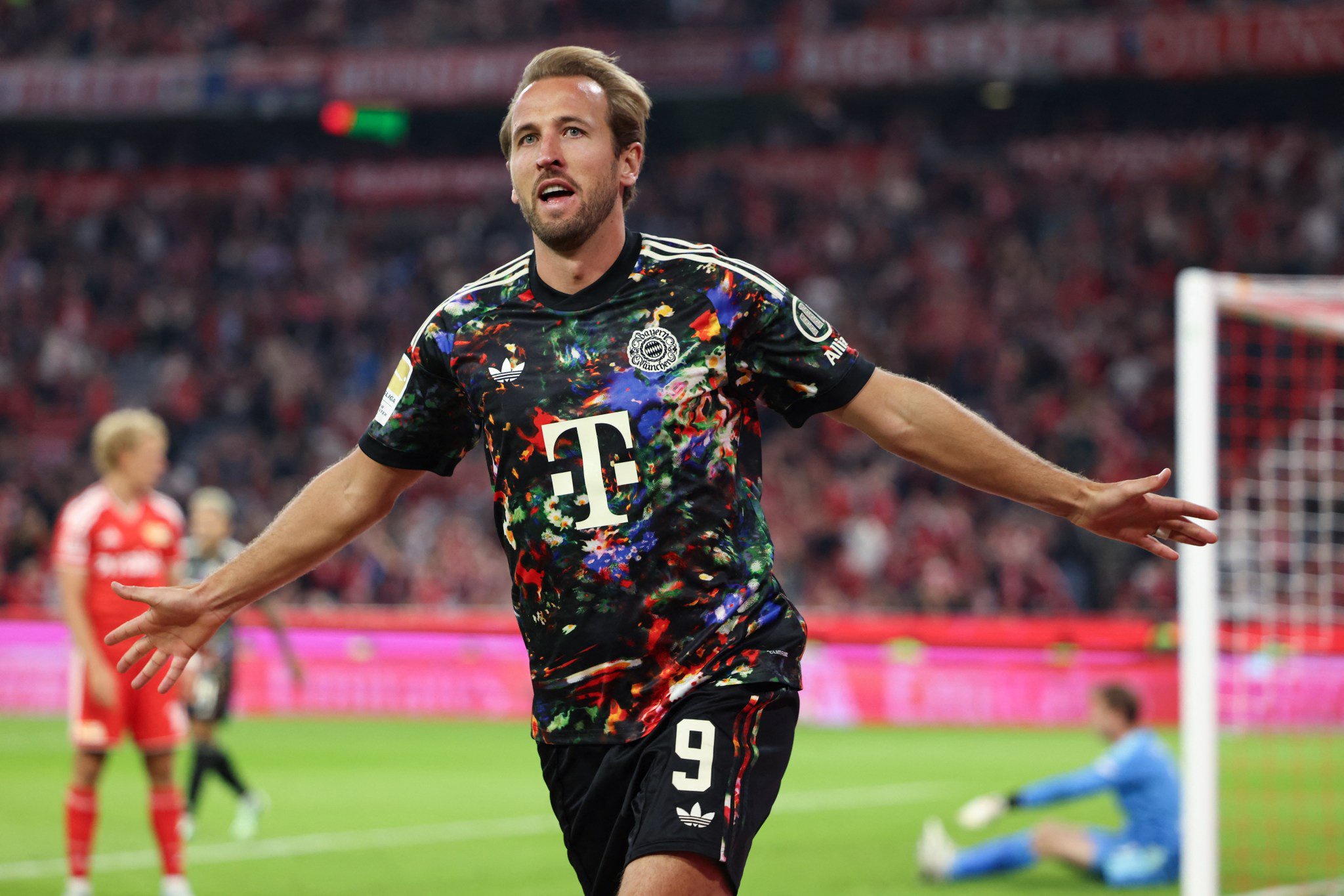 Jogador de futebol Harry Kane, com camisa preta estampada e shorts pretos com o número 9, comemora um gol de braços abertos no campo, com torcida ao fundo