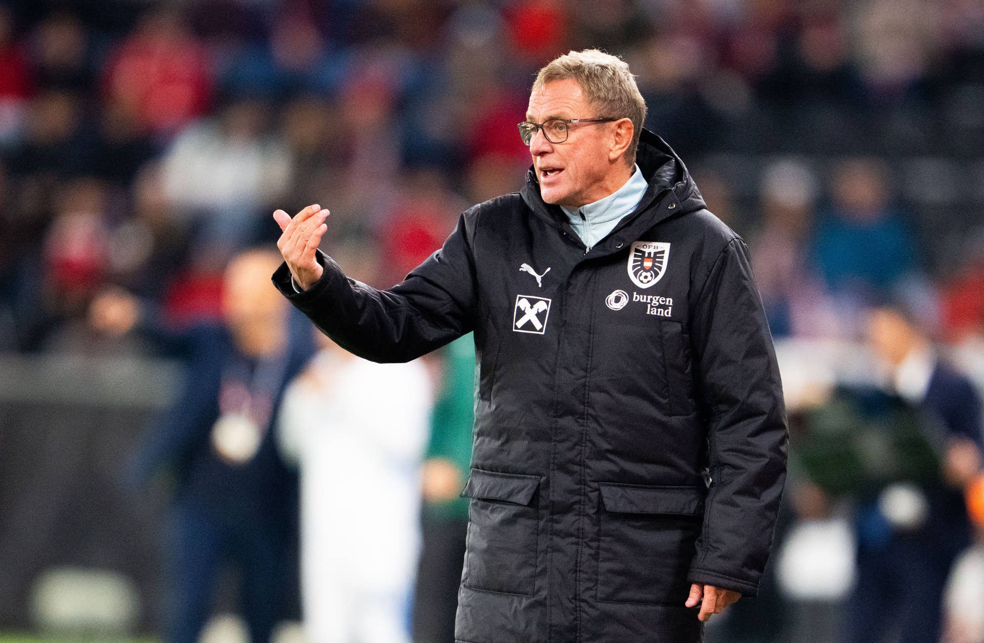 Ein Fußballtrainer in dunkler Jacke gestikuliert am Spielfeldrand während eines Spiels.