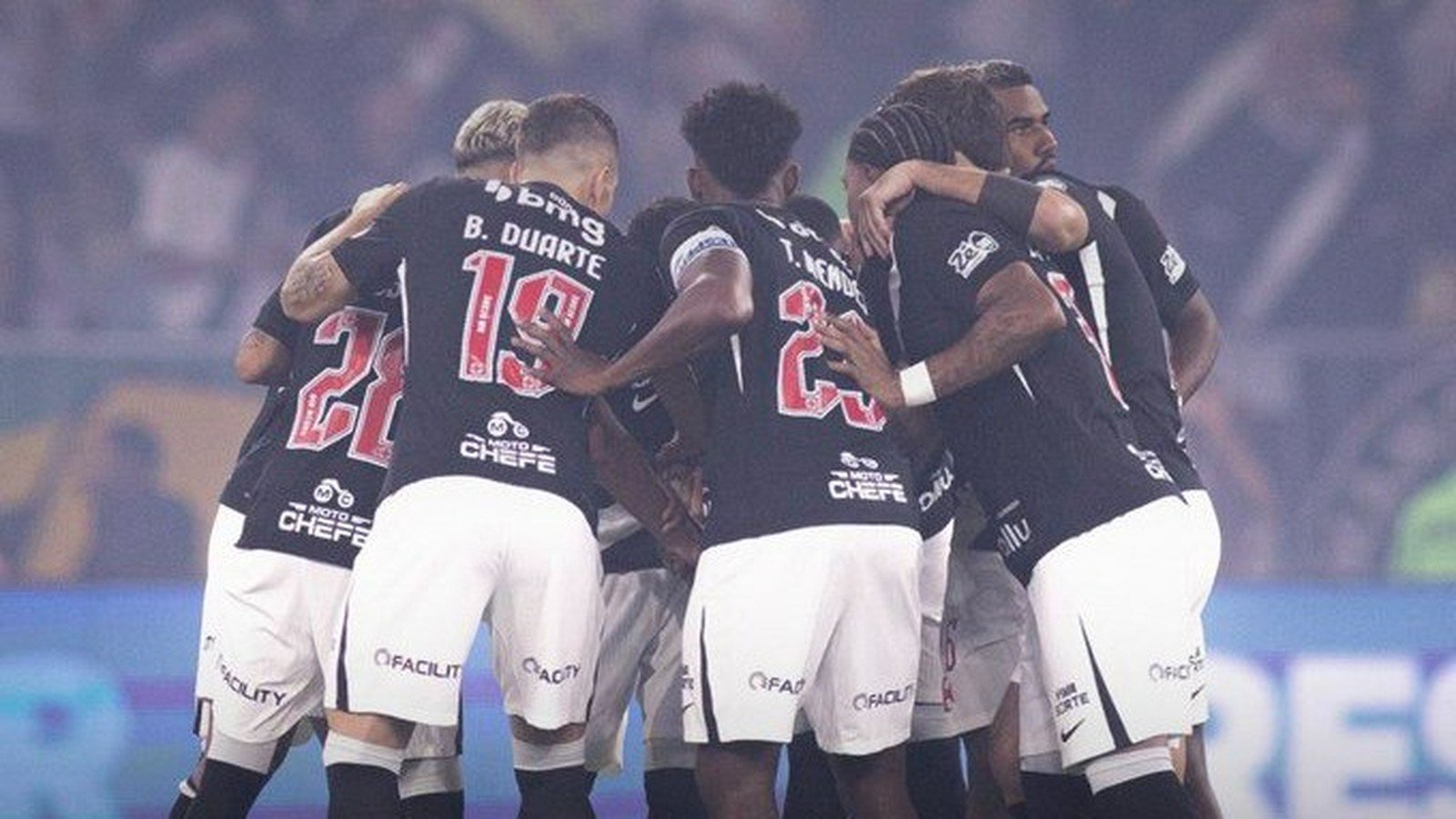 Jogadores de futebol de uniforme preto e branco com logos 'Moto Chefe' e 'Facility' huddle em campo