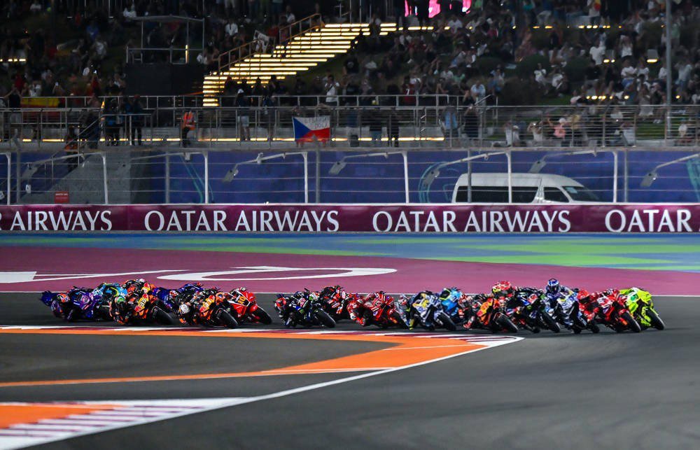 MotoGP riders race in a tight pack past Qatar Airways banners at the Losail circuit during the Qatar Grand Prix at night.