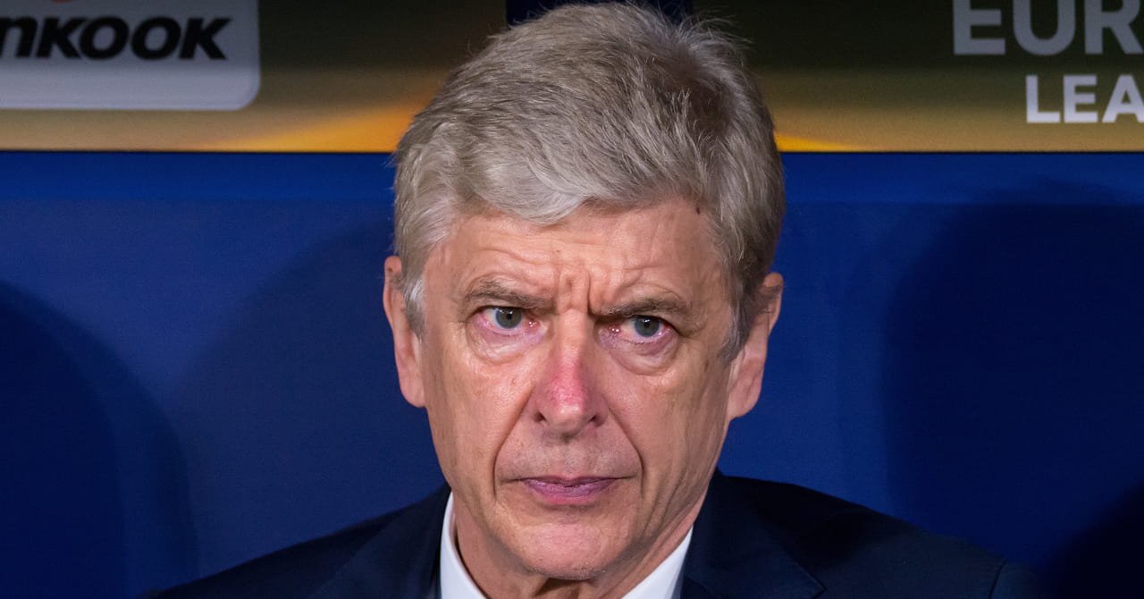 A photo of football manager and legendary Arsenal Football Club head coach Arsene Wenger sitting in a technical area looking stern. Blue seats are visible in the background alongside signs