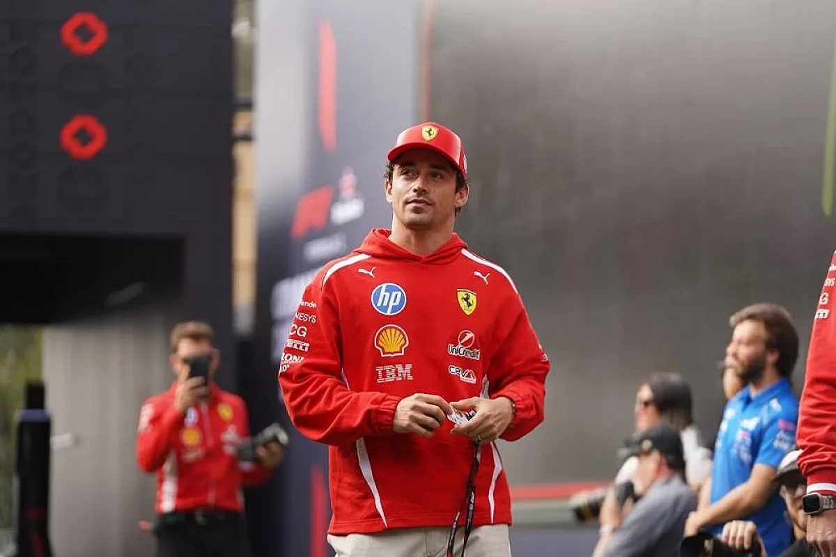Driver in red Ferrari hoodie and cap walking in the F1 paddock, with Shell, HP and IBM sponsor logos visible on his team kit.