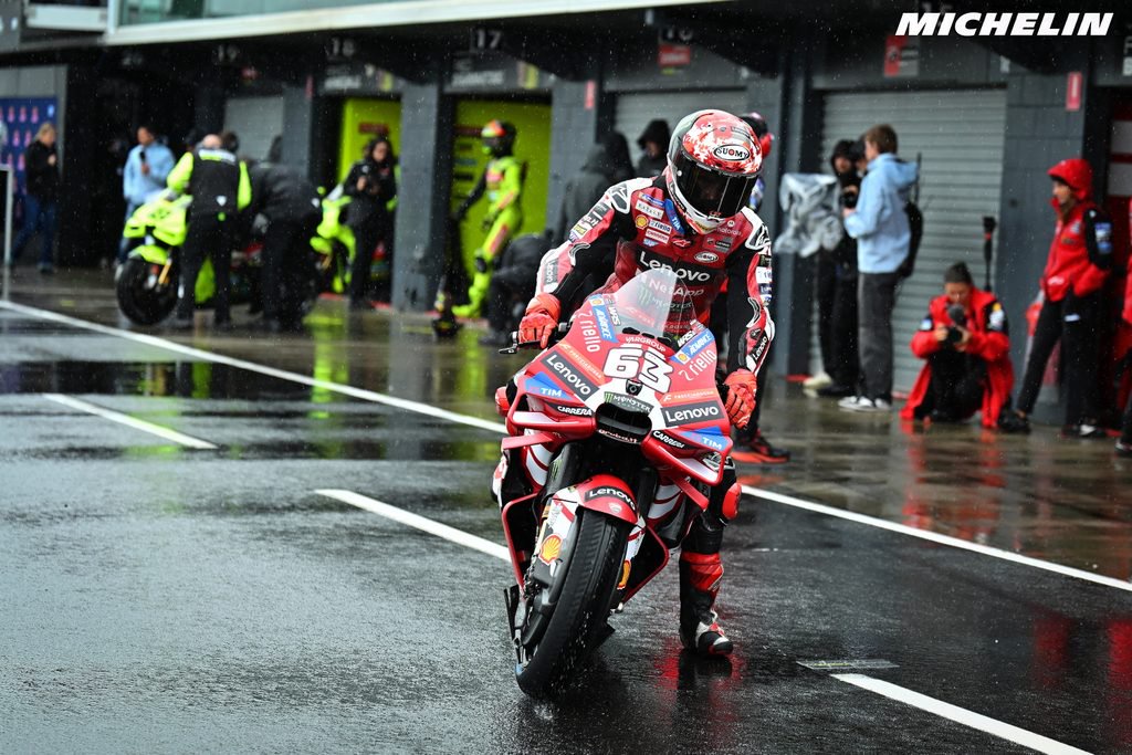 A rider in red and black leathers pushes a red Ducati MotoGP bike through a wet pit lane, race number 63 visible, Lenovo livery.