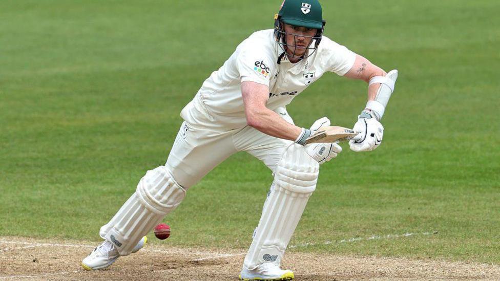 Adam Hose in action for Worcestershire wearing a white cricket outfit while carrying a brown wooden bat on a green pitch with a red ball by his right foot