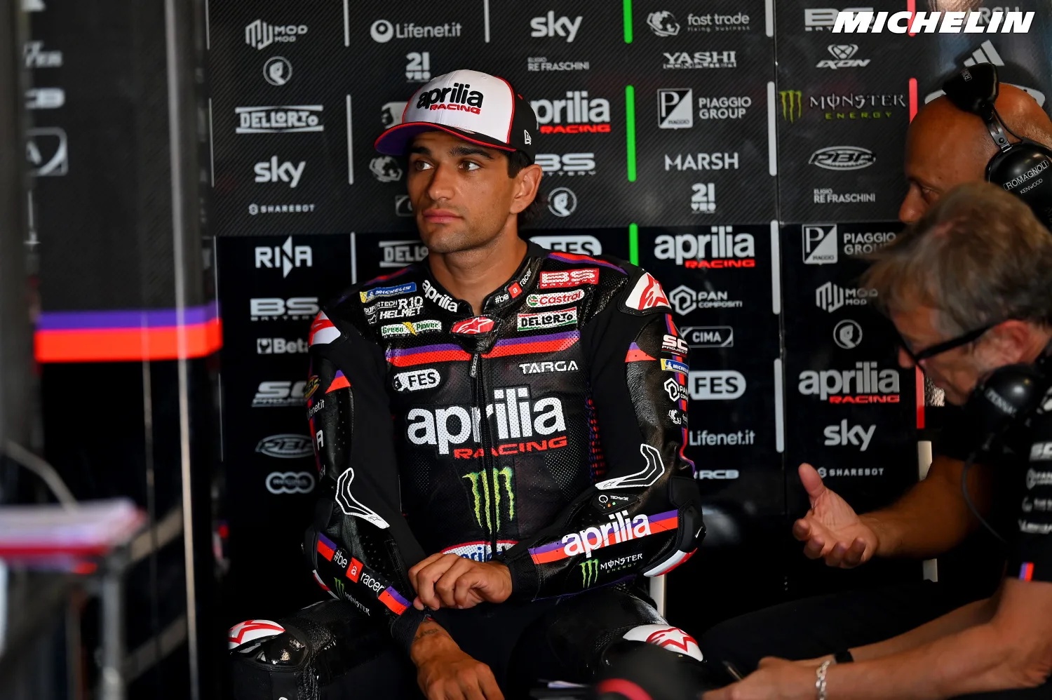Aprilia rider in team leathers and cap seated in the garage beside crew members, with Aprilia Racing and Monster Energy branding behind him (MotoGP)
