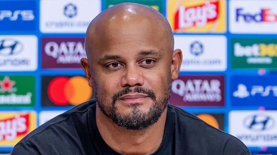 Vincent Kompany mit Glatze und kurzem Bart trägt ein schwarzes Shirt und sitzt vor einer blauen Wand mit Sponsorenlogos.