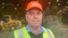 Joseph Doherty stands outside a pile of recycling wearing an orange hard hat and yellow hi-vis jacket