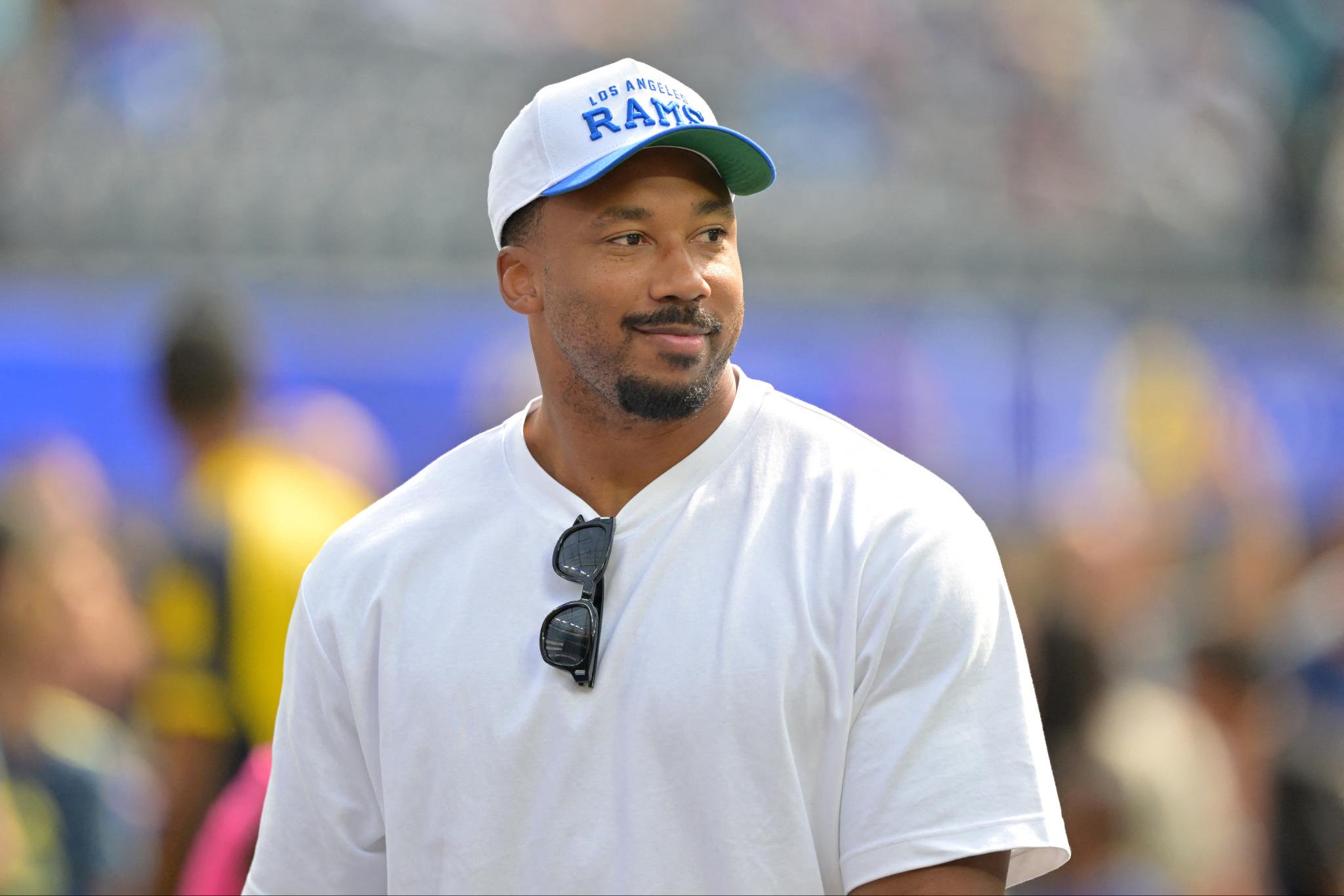 A man wearing a white Los Angeles Rams hat and a white t-shirt stands on the sideline.
