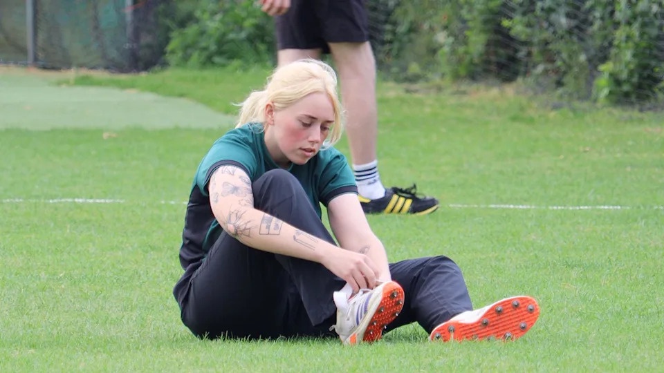 A female cricketer wearing a green top and navy trousers with white shoes. She's sitting on a cricket pitch.