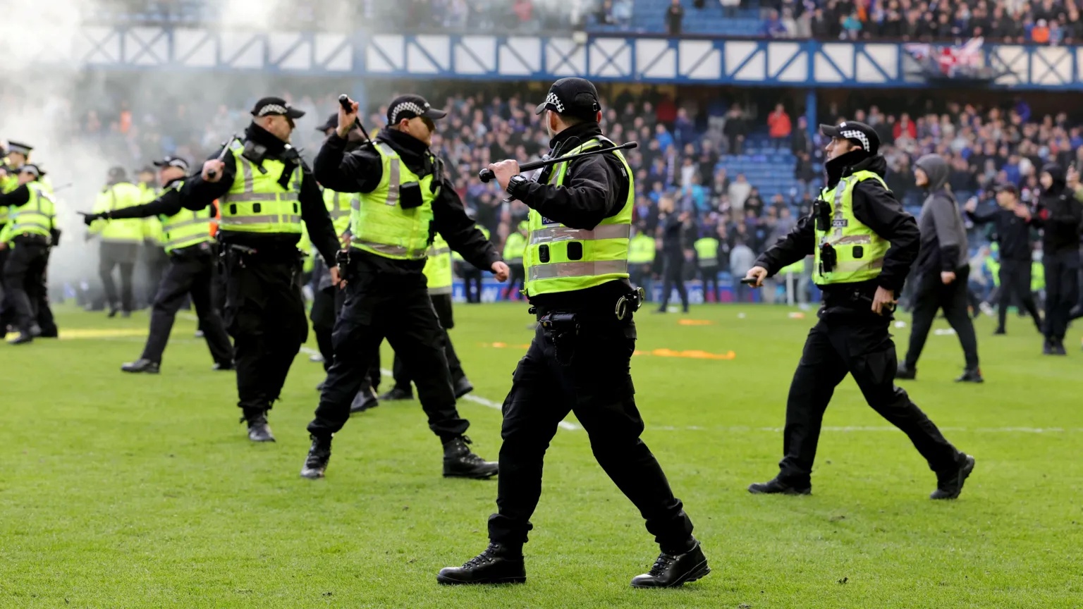 PA Media Police officers wearing black trousers, black jackets, hi-vis vests and black caps holding raised batons on a football pitch. There is smoke in the background. Fans can be seen on the pitch and in the stands.

