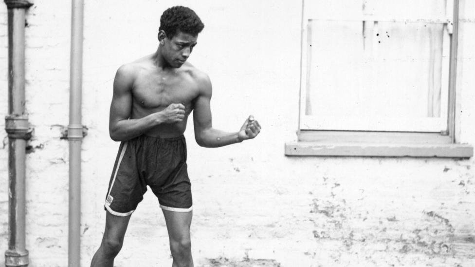 Boxer Len Johnson, shirtless, stands side on in front of a white wall