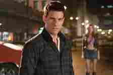 Tom Cruise stands in a dark city street with a woman in the background. Cruise wears a casual plaid shirt