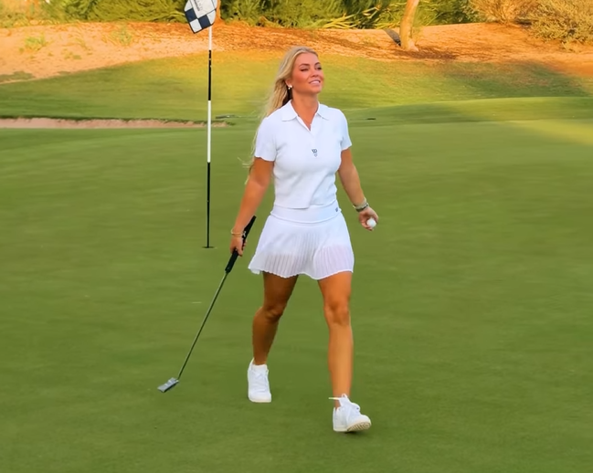 A blonde woman walking on a golf course holding a putter and a golf ball.