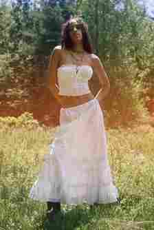 A woman stands in a grassy field, wearing a white strapless crop top and a long white skirt. She has her hands behind her back and looks confidently at the camera