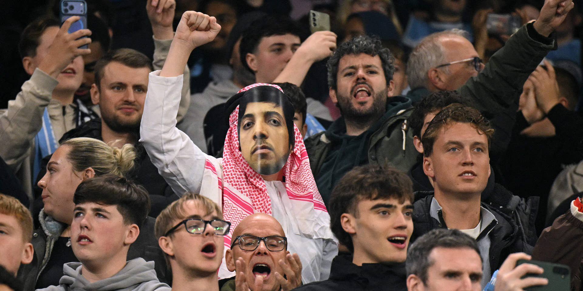 Ein Fan trägt eine Maskerade im Stadion, umgeben von jubelnden Zuschauern bei einem Fußballspiel.