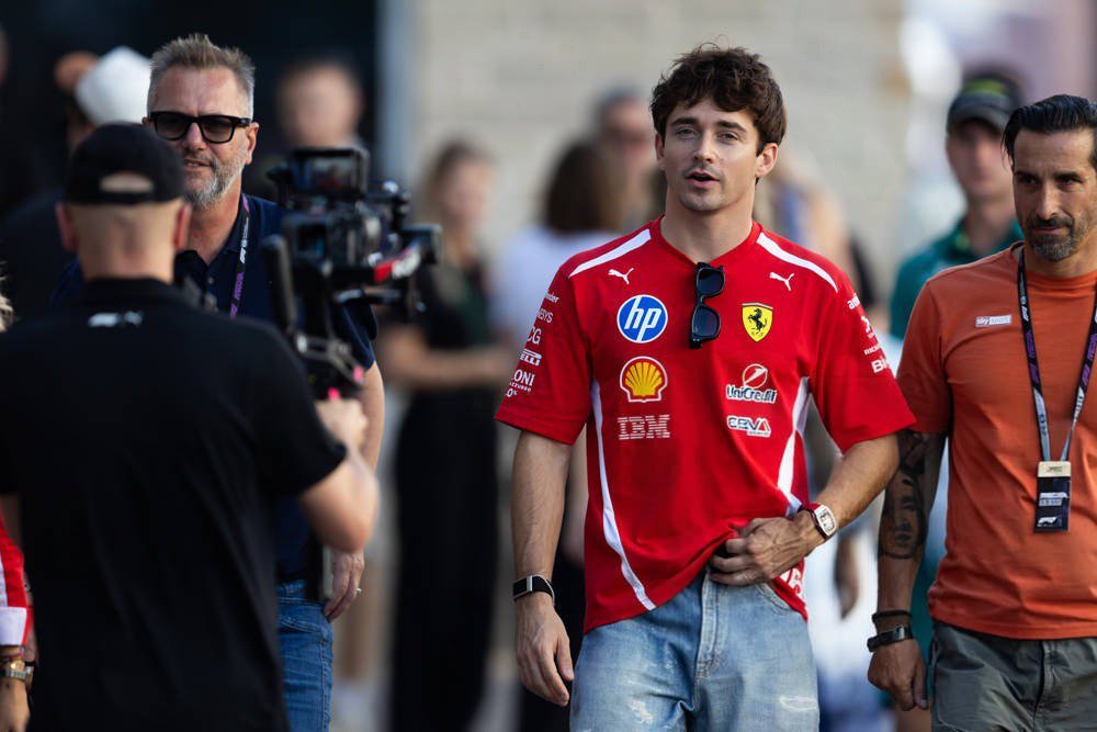 Driver in red Ferrari t-shirt with HP, Shell and IBM sponsor logos walks through a paddock as cameras film nearby, F1 pass visible