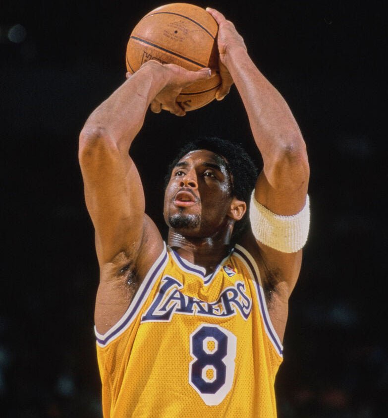 A basketball player in a yellow Lakers jersey, number 8, is shown mid-jump as he makes a shot. He is focused, with both arms extended upwards, and a white wristband on his right arm. The background is dark, highlighting the player.